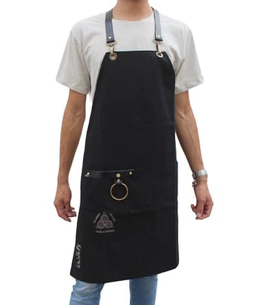 Modelo usando avental de chef na cor preta com a marca Walls impressa em sua superfície. Possui detalhes em metal e apresenta a frase "De Minas para o Mundo"