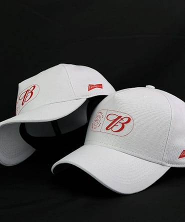 Dois bonés brancos com estampa da Budweiser na lateral e logo na parte frontal. O logo é composto pela letra B em letras decorativas e um símbolo de uma bola de futebol.